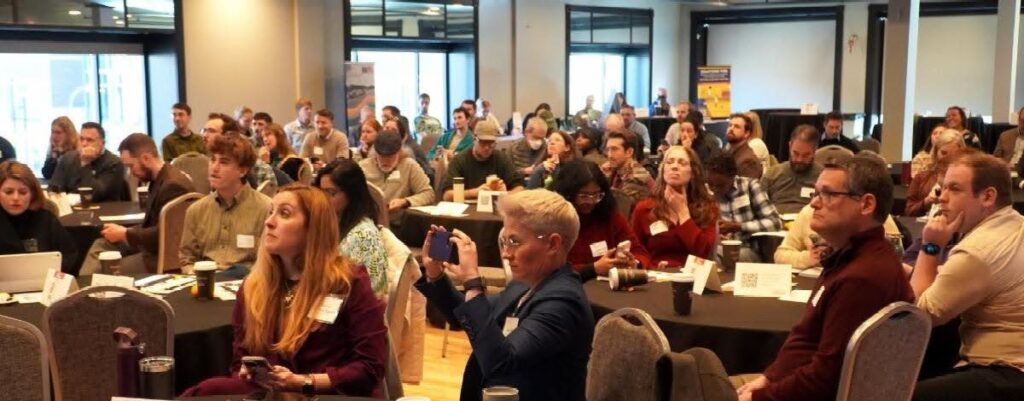 Many people sit at tables and look to the left of the screen at a presenter not in the picture. 