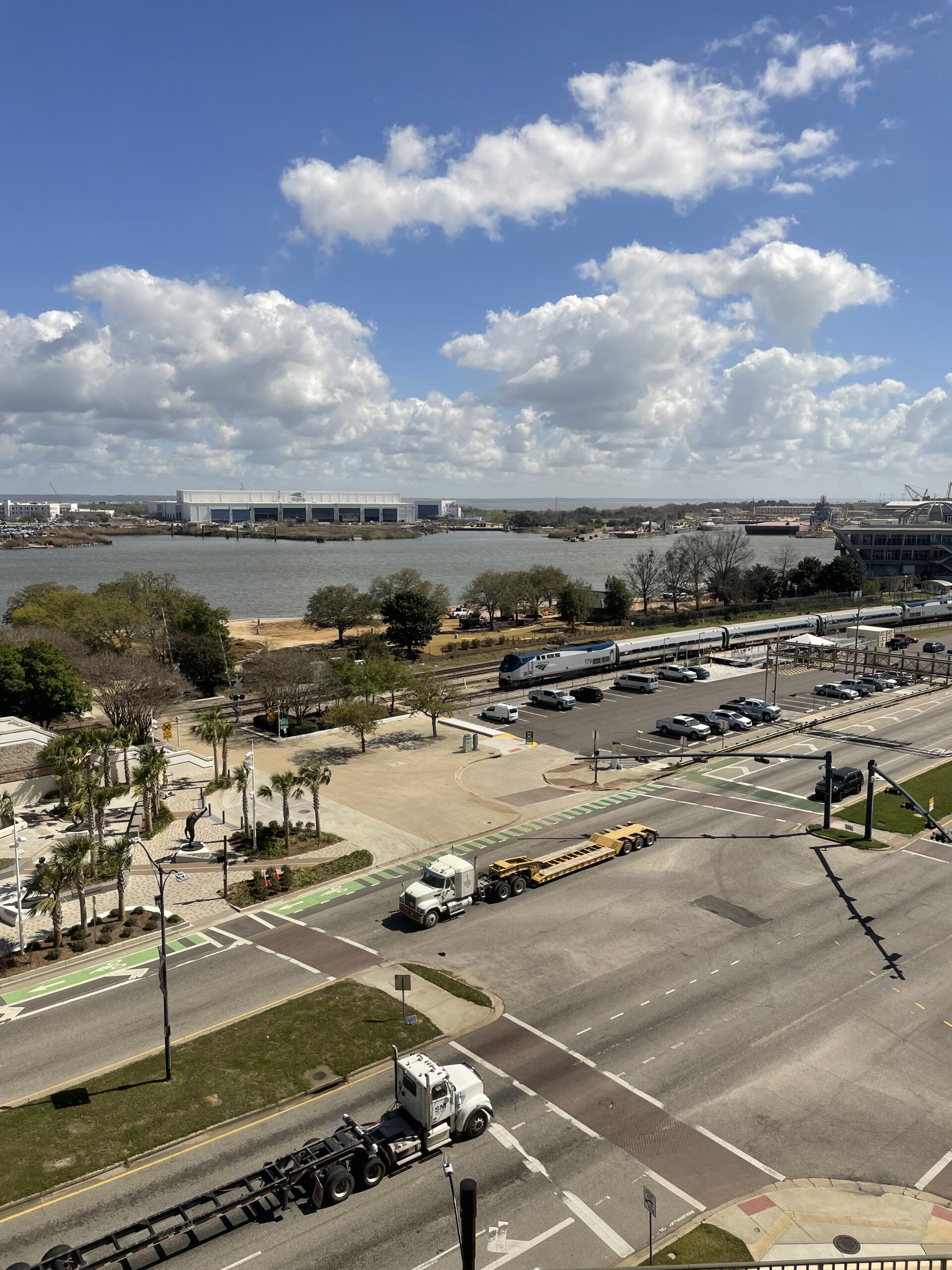 Aerial view of two trucks driving in opposite directions along a street with train tracks and an amtrak train in the background, and a wide river further in the background