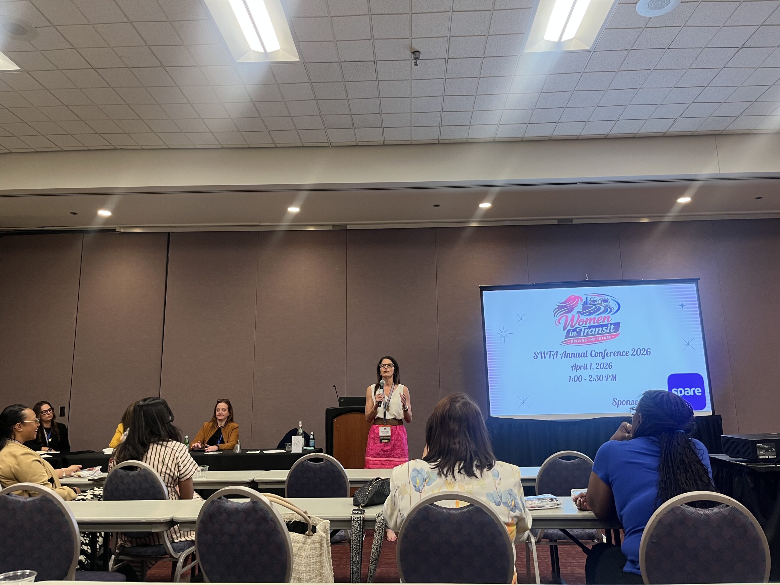 Kendra McGeady standing in front of the room, introducing the Women in Transit panel in a packed room.  