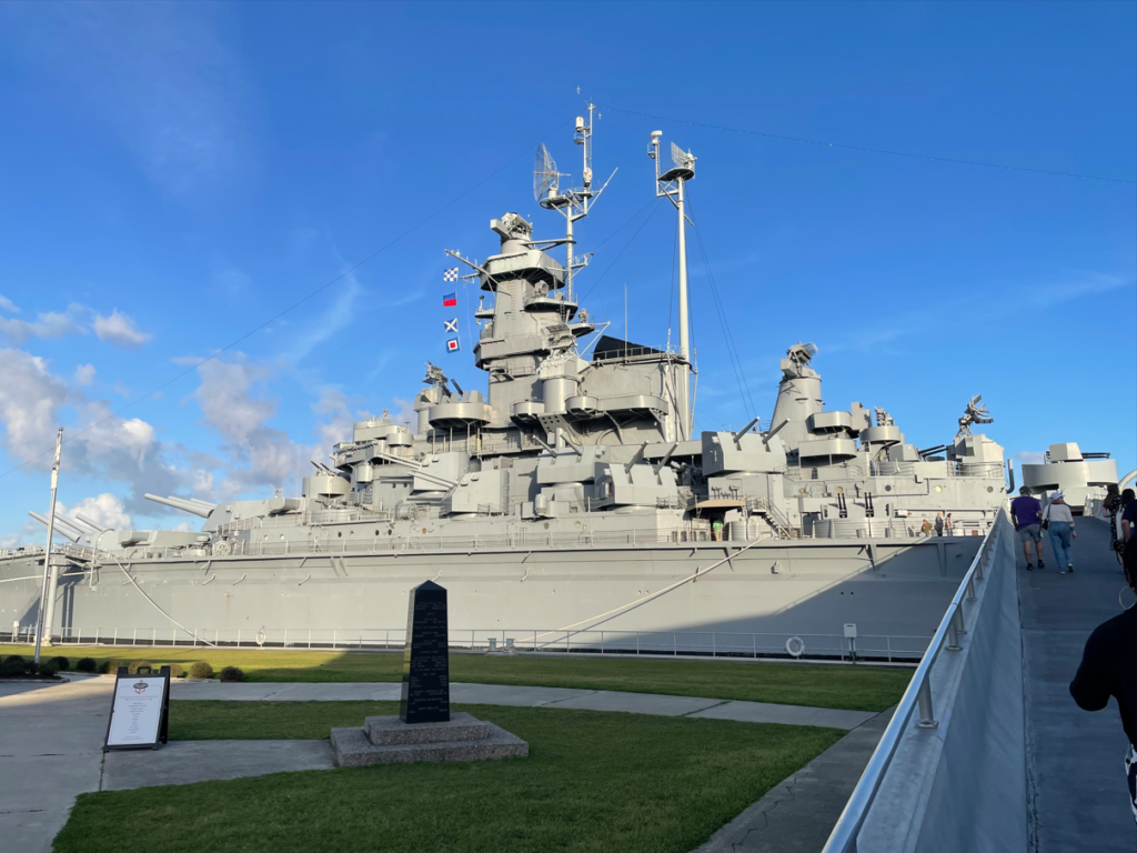 People walk up a ramp on the right side of the image to a large battleship
