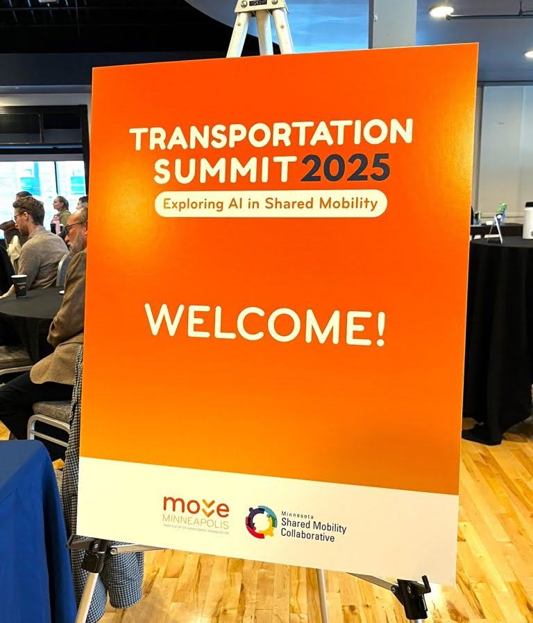 An orange sign with the words “Transportation Summit 2025, Exploring AI in Shared Mobility” and “Welcome!” with the logos of Move MN and MN Shared Mobility Collaborative. 