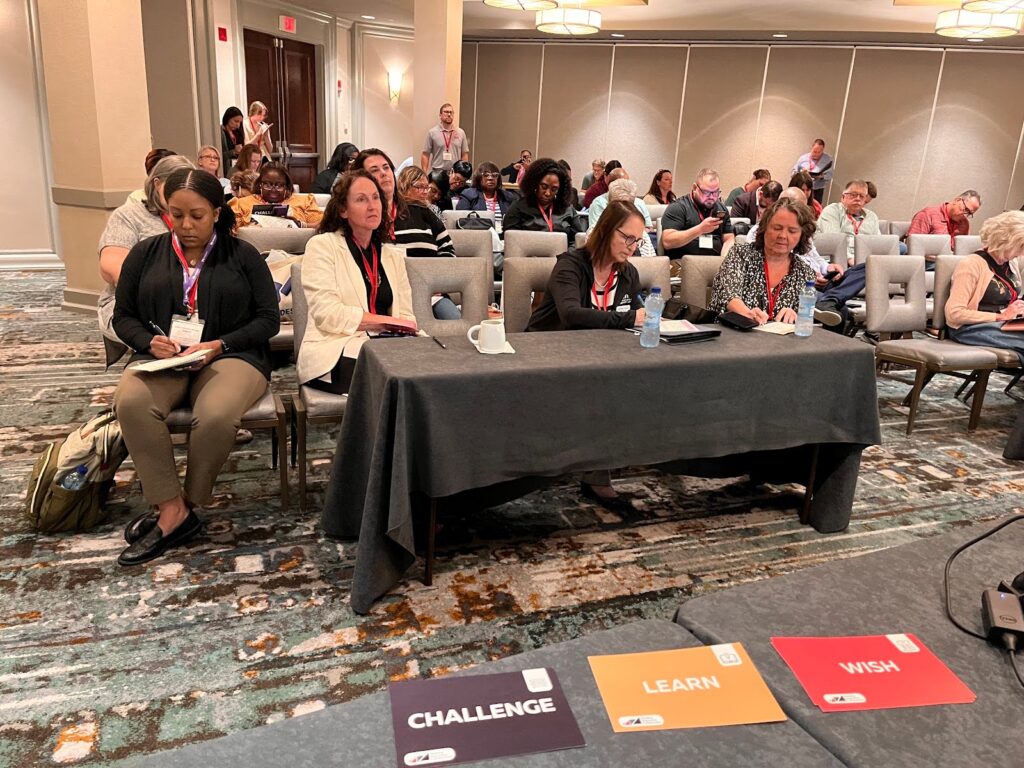 many people sit and write in the background. in the foreground three notecards are on a table: a purple notecard with "CHALLENGE" a yellow notecard with "LEARN" and a red notecard with "WISH"