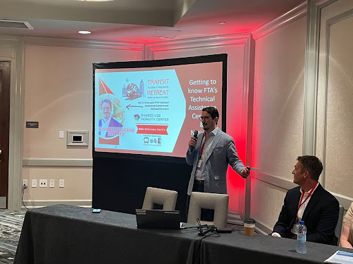 A man stands in front of a projector with a microphone. Another man sits to his left.