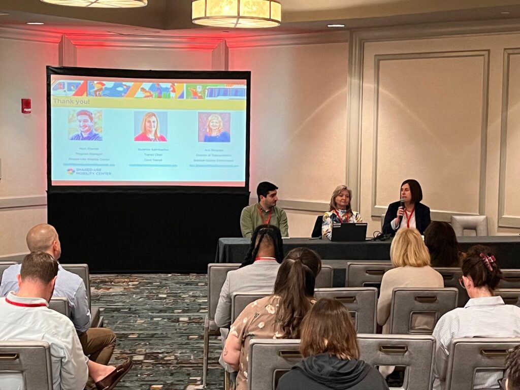 Three people sit at the front of a room next to a projector screen. The woman on the right is holding a microphone. Several audience members look forward at them.